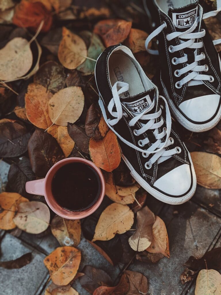 Black Converse sneakers and a coffee cup on autumn leaves, evoking a cozy fall mood.