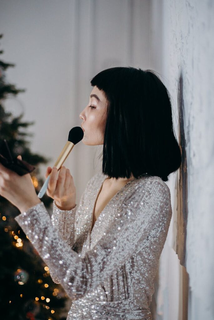 A woman in a silver dress applies makeup indoors beside a decorated Christmas tree.