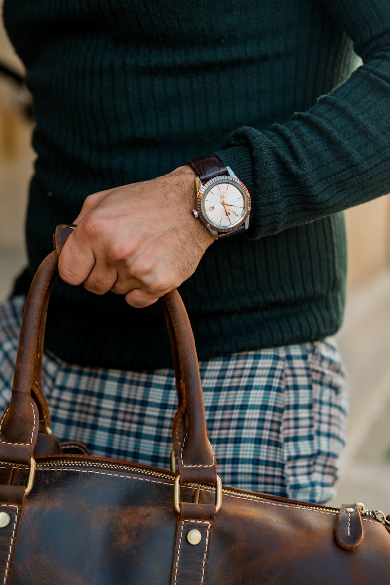 Close-up shot of a person in a green sweater holding a leather bag and wearing a wristwatch.