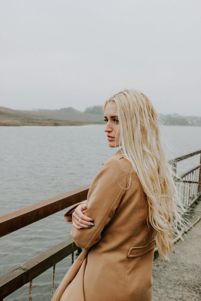 Profile of a woman with long blonde hair in a coat standing on a bridge overlooking a serene lake.