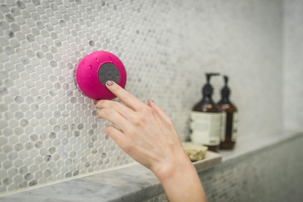 A person adjusting a pink waterproof bluetooth speaker in a tiled bathroom setting.