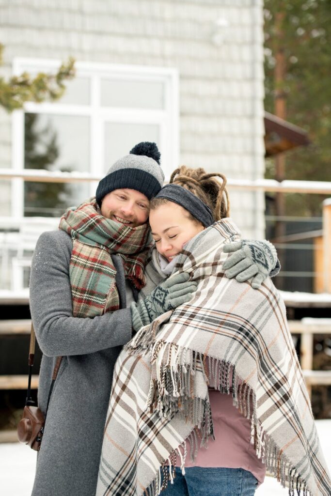 A couple bundled in scarves and coats embracing each other warmly outside a house in winter.