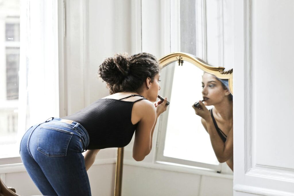 A woman applies lipstick in front of a mirror with a reflection, emphasizing casual beauty indoors.