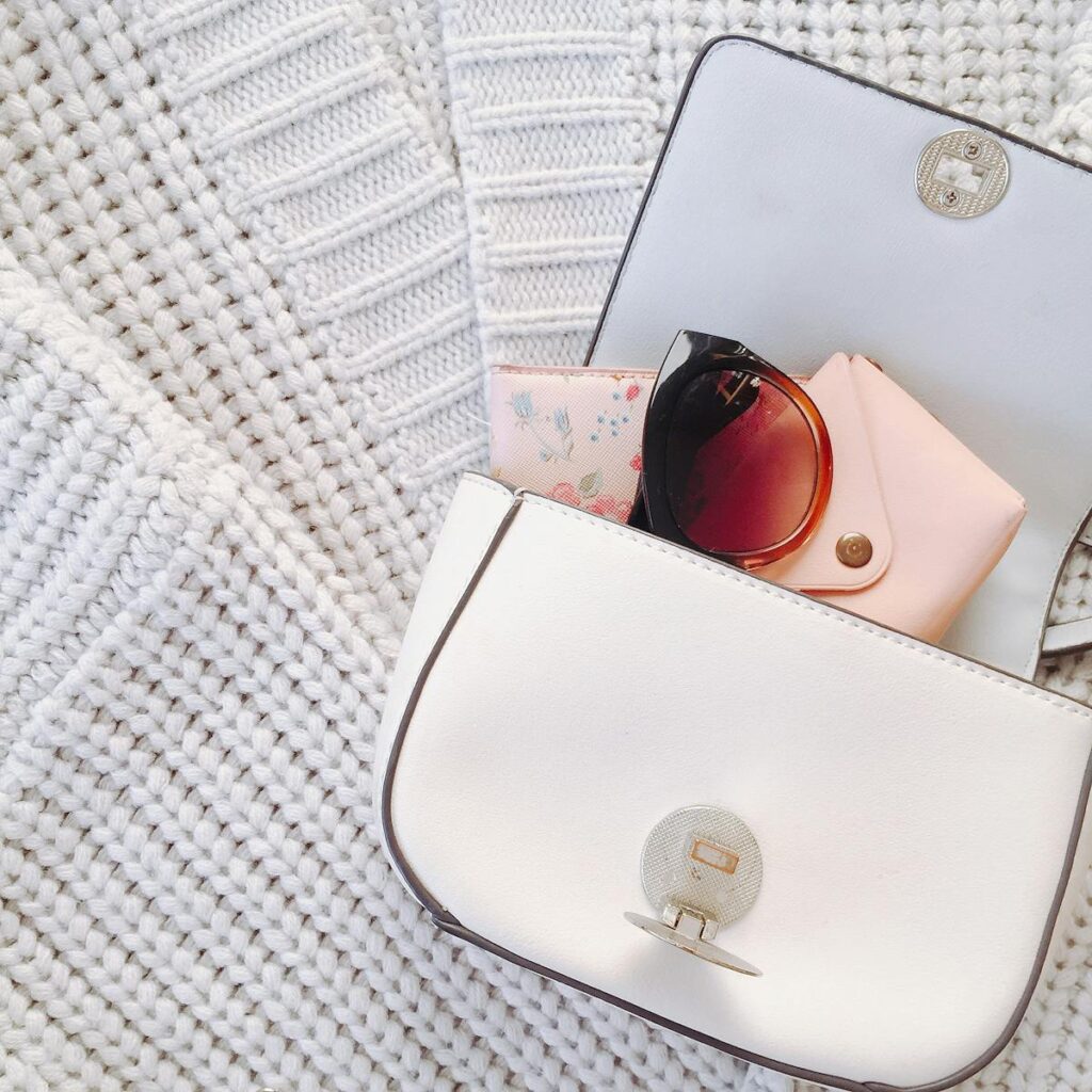 Chic white handbag with sunglasses and accessories on knit sweater, showcasing modern fashion style.
