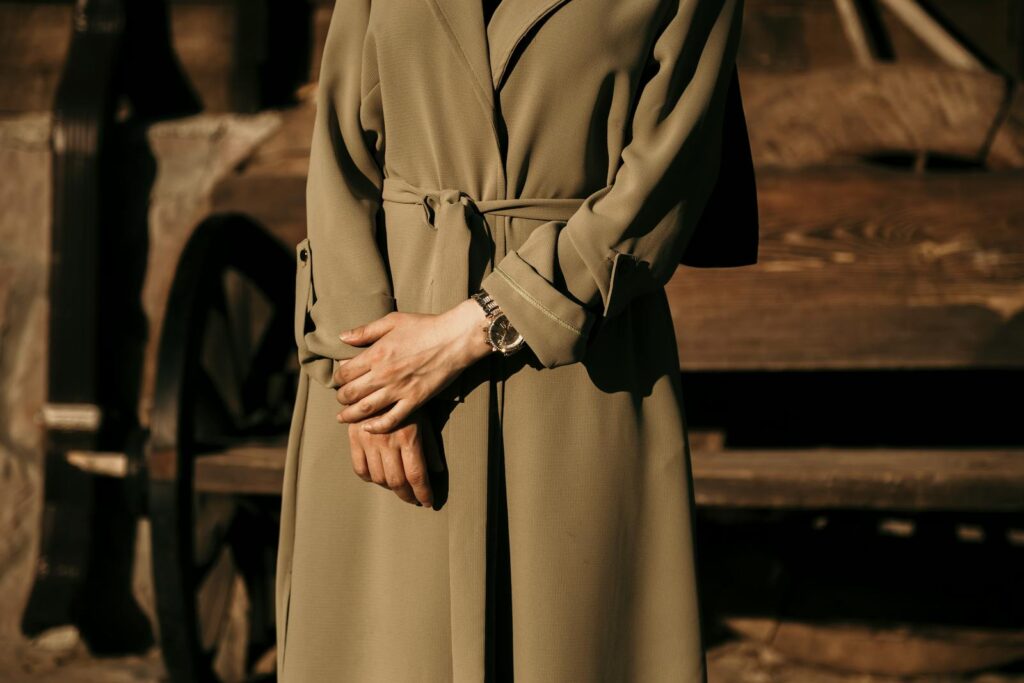 A stylish woman stands outdoors wearing a khaki trench coat and wristwatch, capturing fashion in a rustic setting.