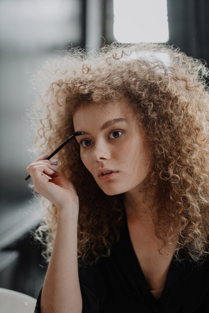 A young woman with curly hair applying makeup, focusing on her eyebrows.