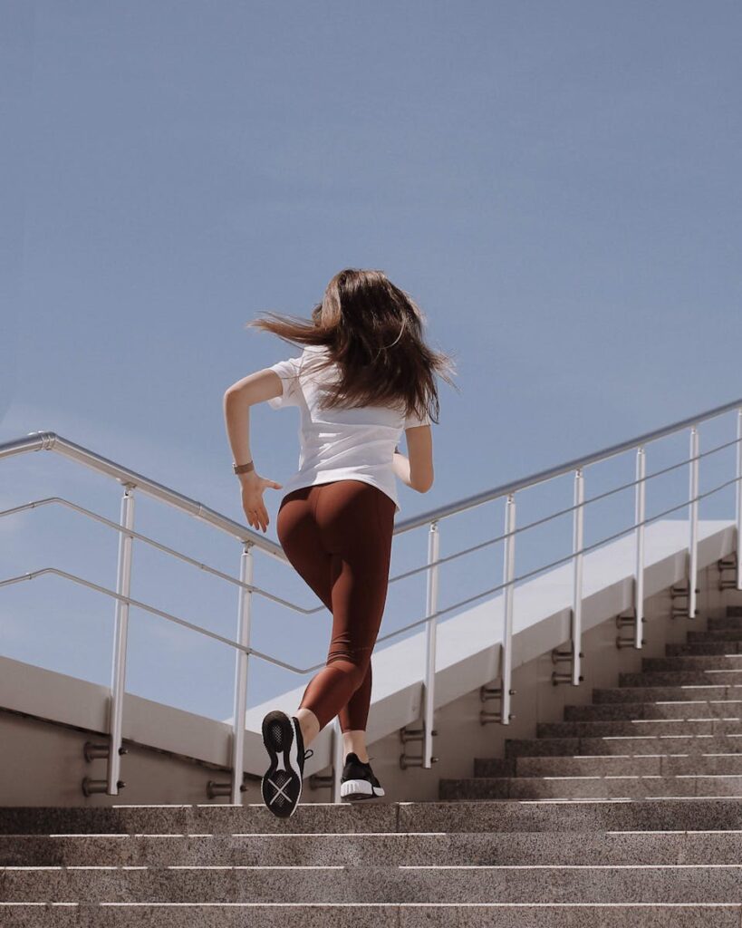 Fit woman in workout attire running up stairs on a sunny day, showcasing active lifestyle.