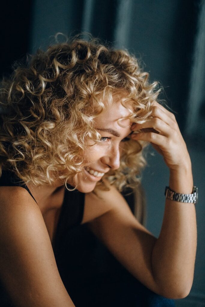 A woman with curly blonde hair and a joyful expression smiles indoors, exuding happiness.