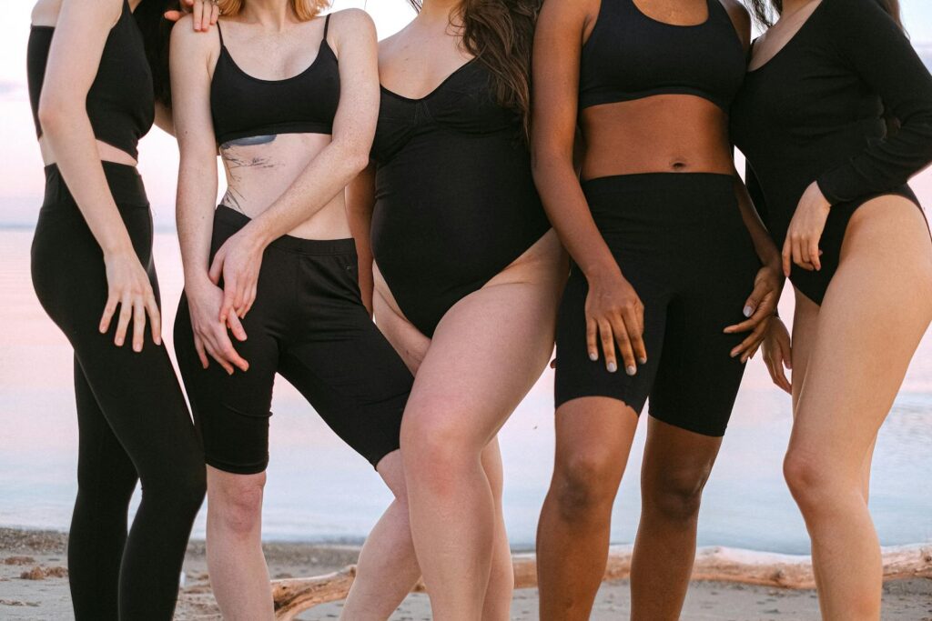 A diverse group of women posing together on a sandy beach with the sea in the background.