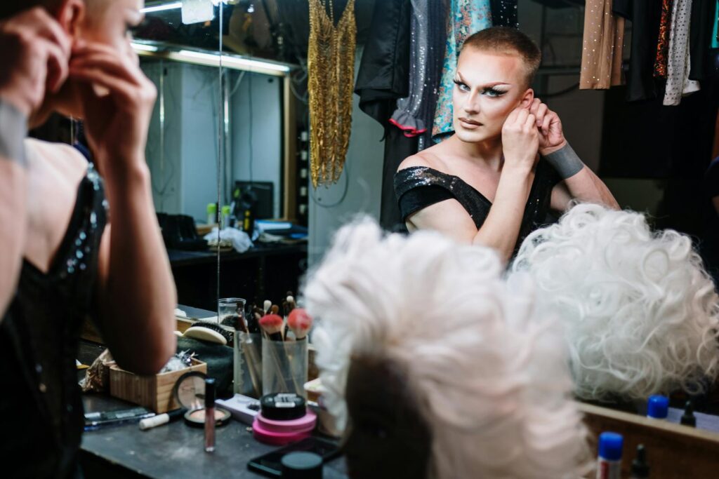 A drag performer gets ready in a dressing room, applying makeup and adjusting earrings.