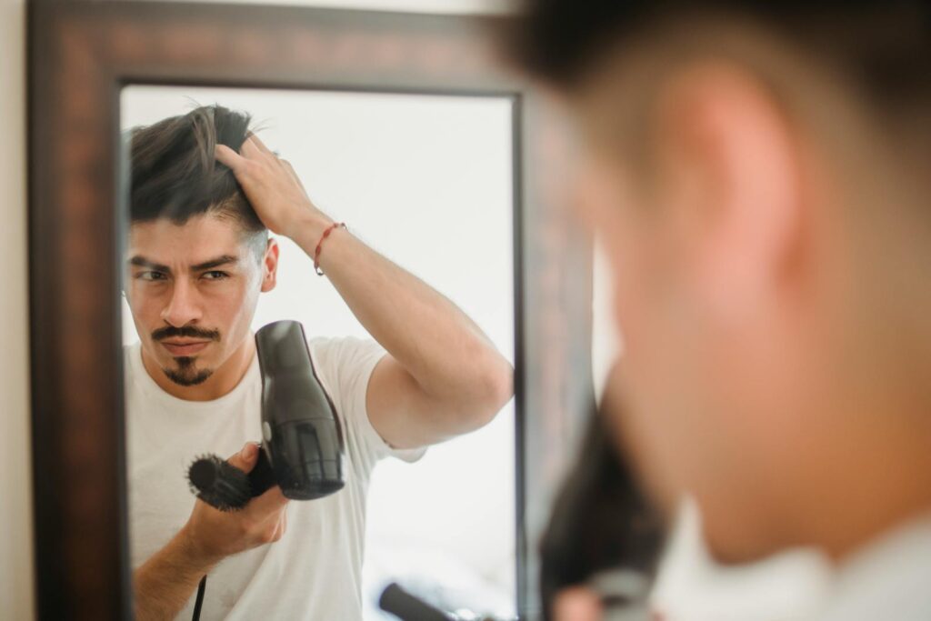 Adult man styles his hair using a hairdryer while looking at his reflection, capturing a modern grooming routine.