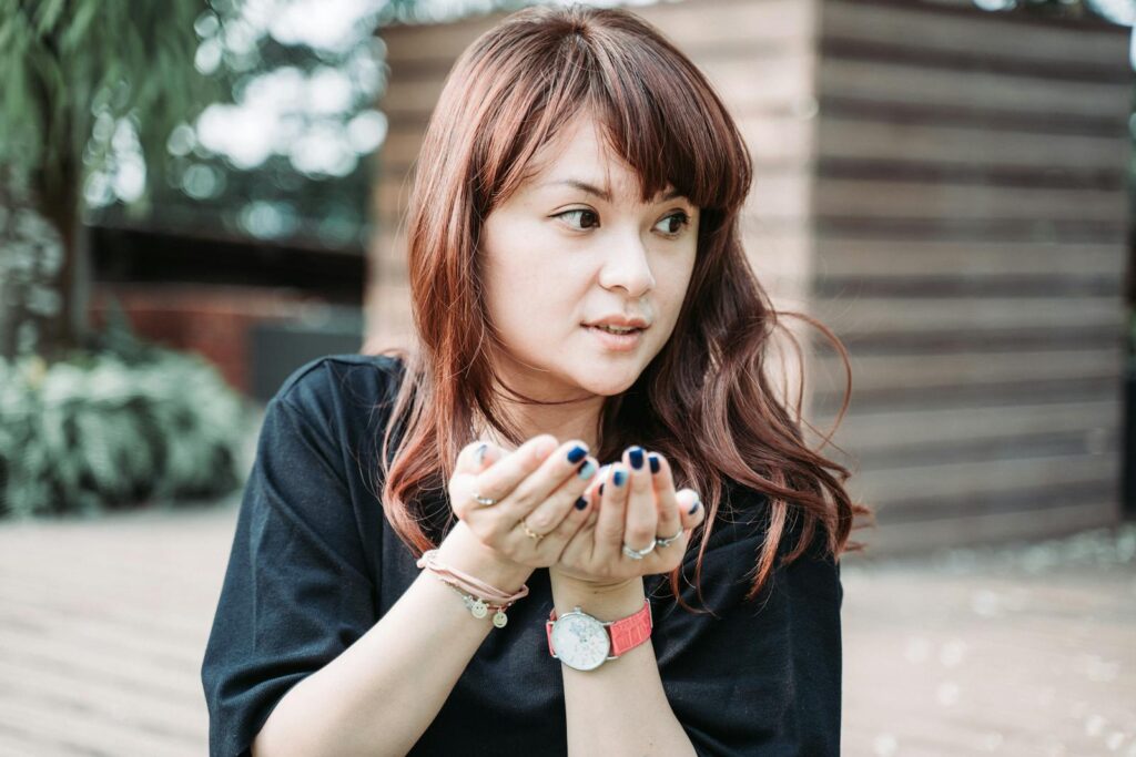 An Asian woman with bangs and casual attire holds her hands out in an outdoor setting.
