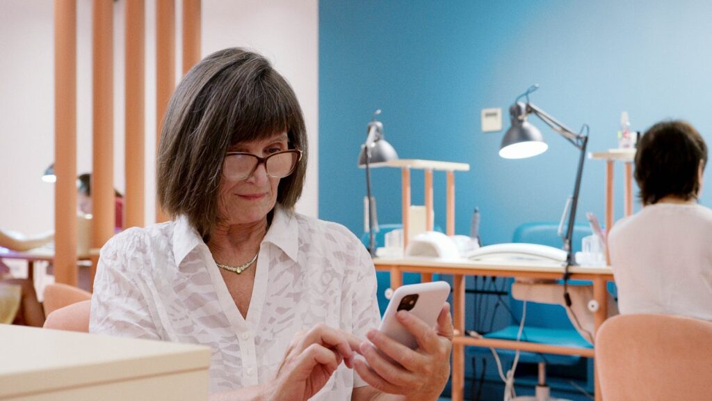 Elderly woman seated in a salon using a smartphone, surrounded by salon equipment.