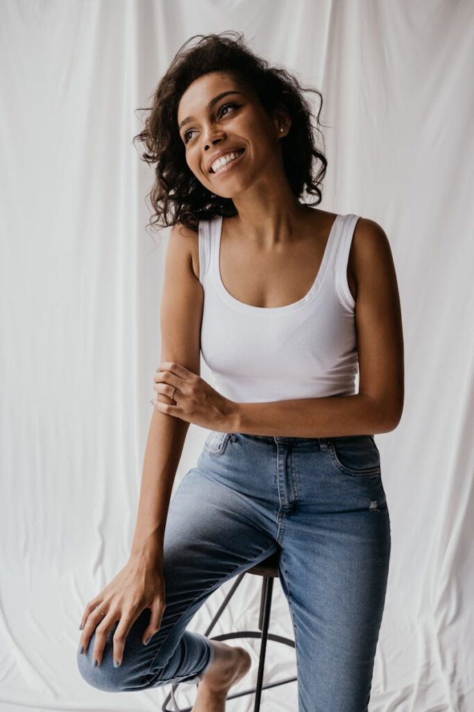 Smiling woman in white tank top and denim jeans sitting indoors on stool.