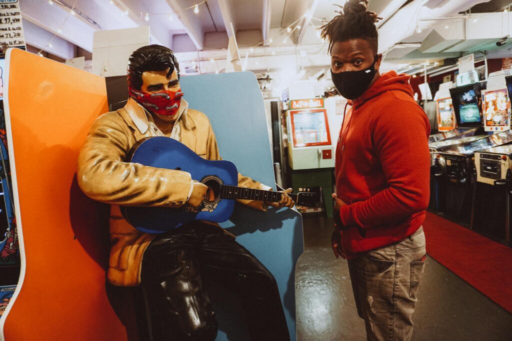 Man in mask stands by Elvis Presley statue with guitar in vintage arcade.