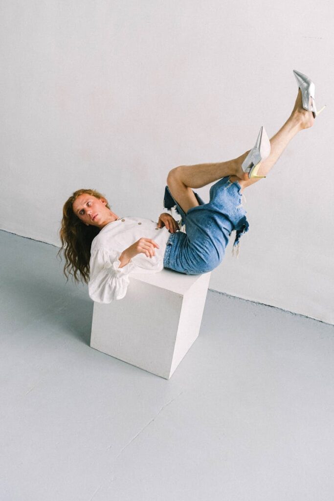 A model in denim and heels strikes a dynamic pose on a cube in a studio setting.