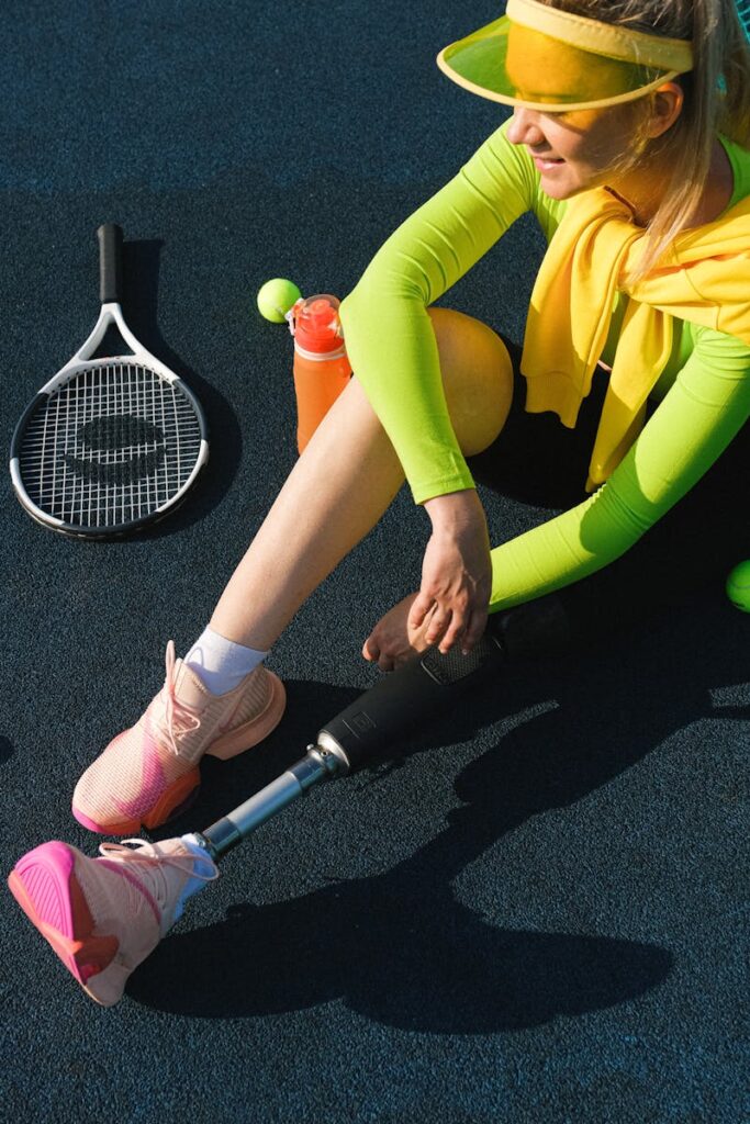 Woman with prosthetic leg sitting on court with tennis gear, exuding vibrant athleticism in neon attire.