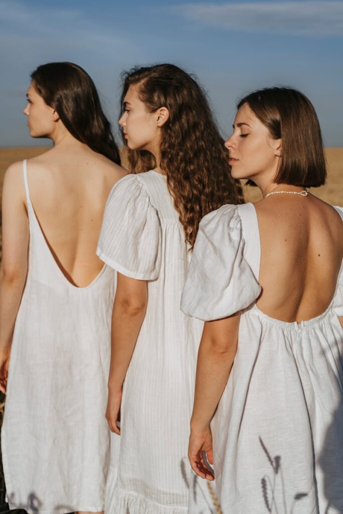 Three women in white dresses standing outdoors with eyes closed, embracing nature's serenity.