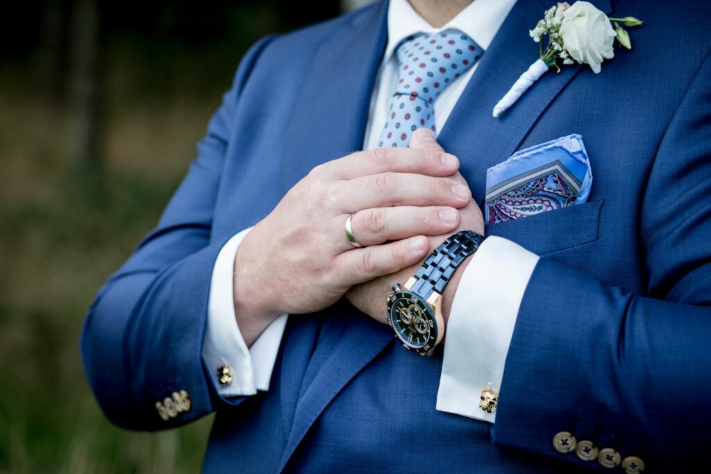 Close-up of a groom in a navy blue suit, wearing a wedding ring and white rose boutonniere.