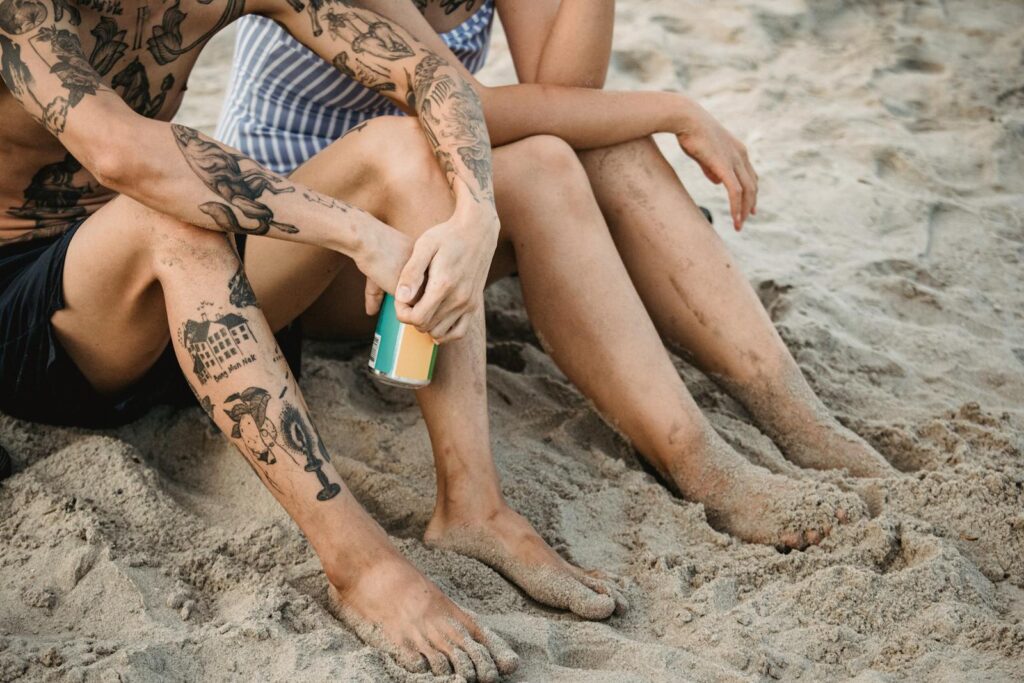 Tattooed couple sitting barefoot on a sandy beach, enjoying a summer day.