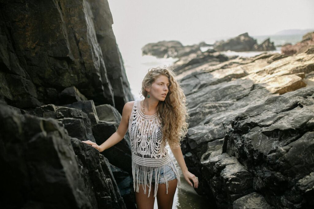 A woman in bohemian attire poses among rugged rocks by the sea, showcasing natural beauty.