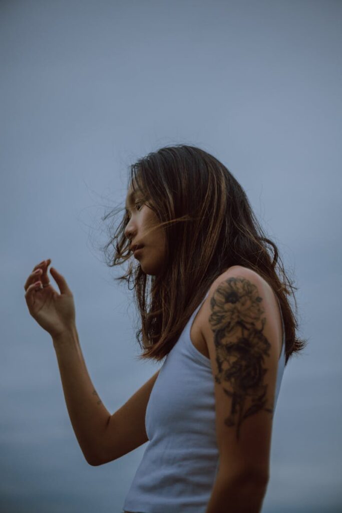 Portrait of a woman with a floral tattoo on her arm, wearing a white tank top against a moody sky.