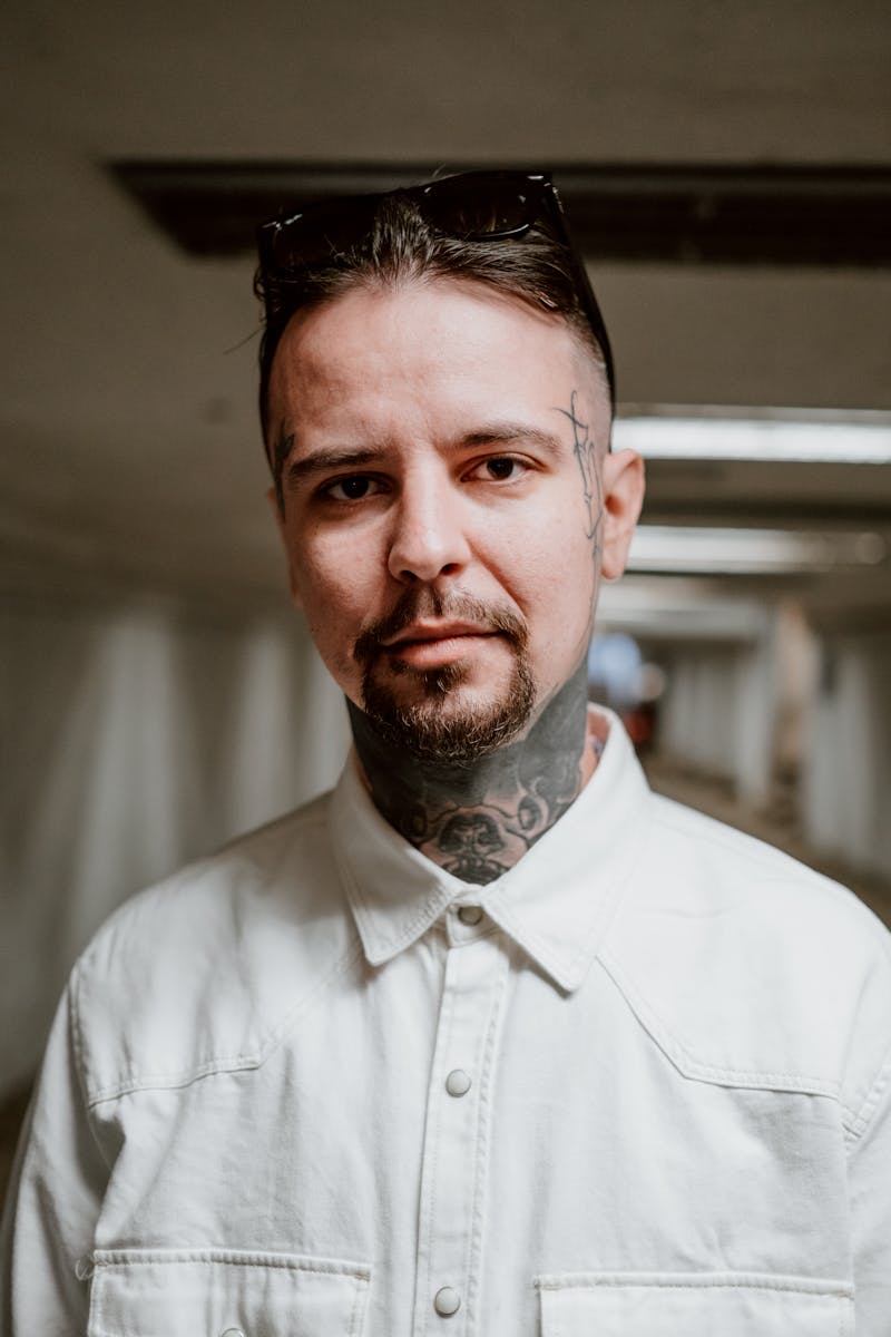Close-up portrait of a stylish, tattooed man wearing a white shirt, standing in an indoor corridor.