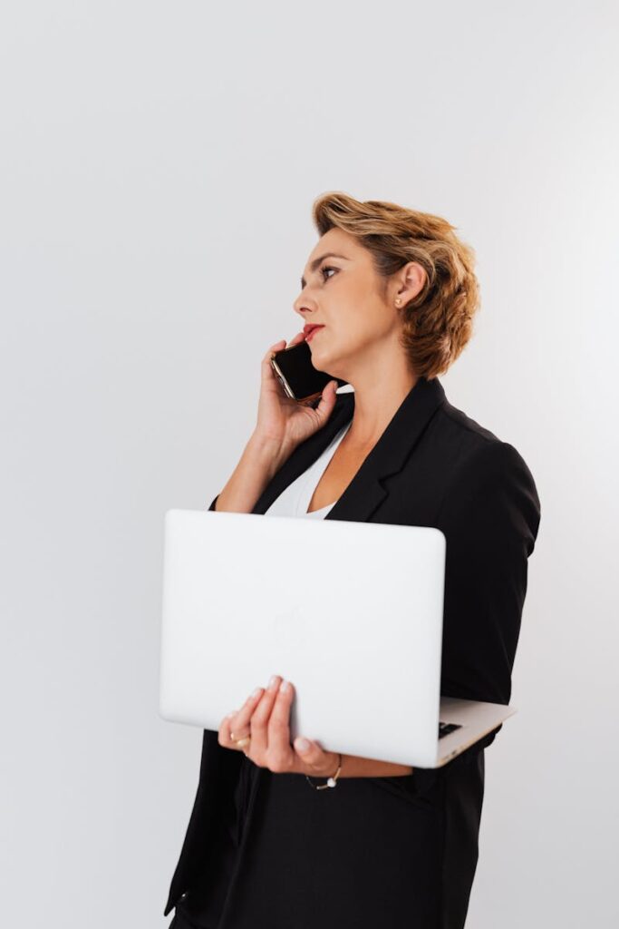 Focused businesswoman in black blazer holds a laptop while talking on the phone.