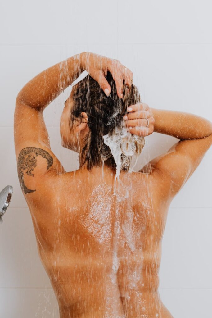 Rear view of a woman washing her hair in the shower, showcasing a tattoo and emphasizing hygiene.