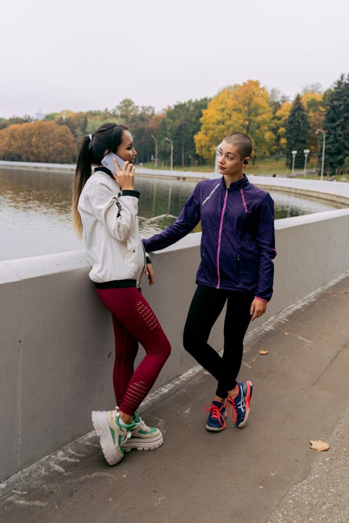 Two women in activewear talking by the lake during a fall day. One is on her phone.