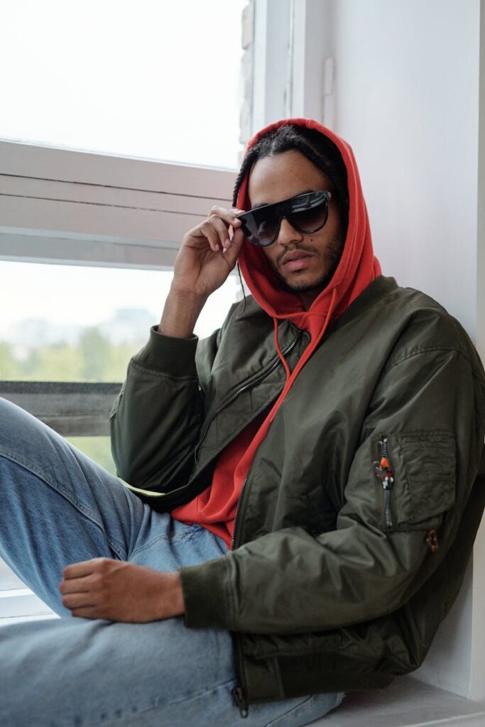 Portrait of a stylish man wearing a hoodie and sunglasses, sitting by a window in streetwear fashion.
