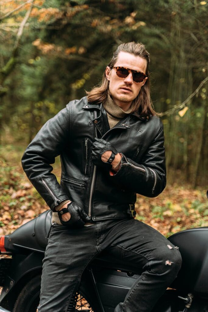 A stylish biker wearing sunglasses and leather jacket sits confidently on a motorcycle in a forest setting.