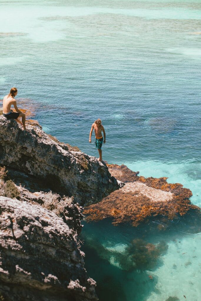 Two men preparing to cliff dive into clear turquoise ocean waters, enjoying a summer adventure.