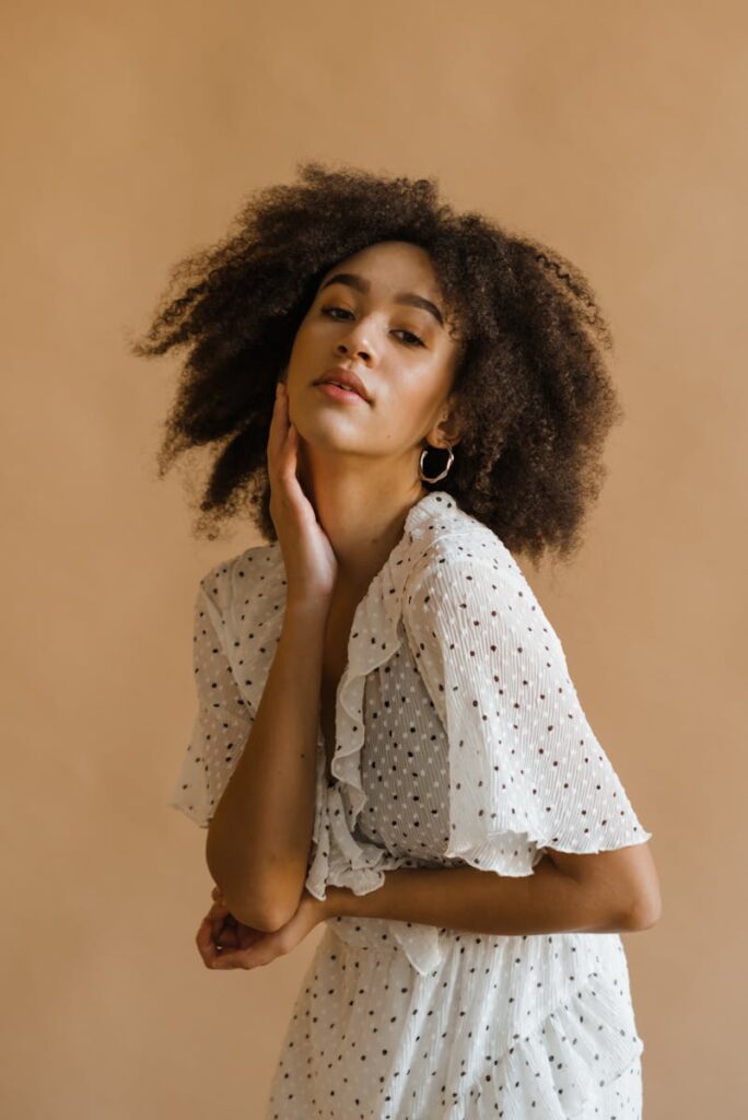 Side view of confident young black female model with Afro hairstyle in stylish dotted dress touching face and looking at camera against beige background