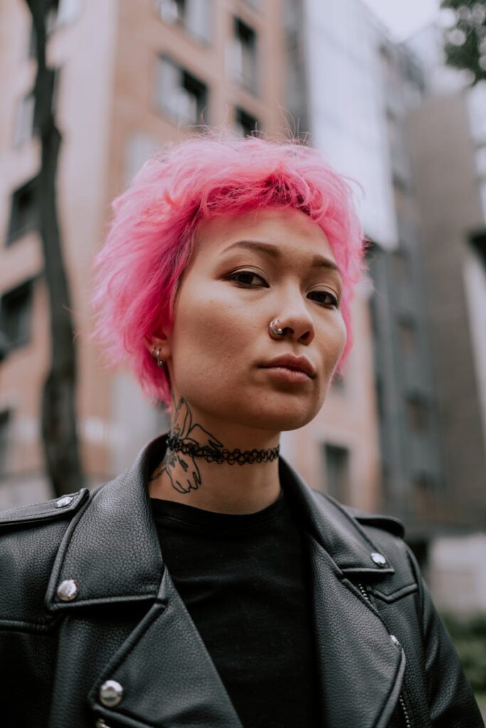 Stylish pink-haired woman with leather jacket and tattoo poses confidently outdoors.
