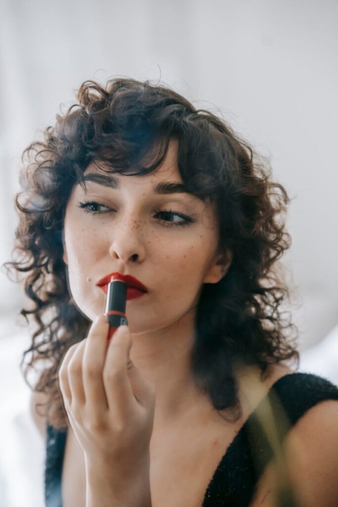 Content confident young female with makeup on face looking at mirror while spreading red lipstick on lips