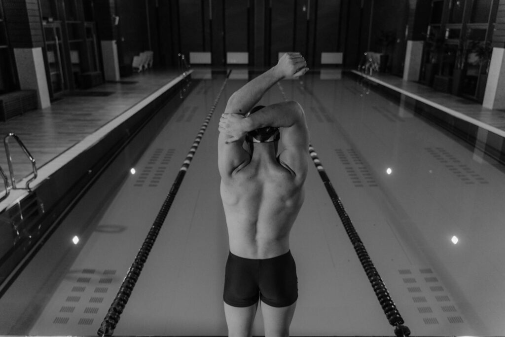 Black and white photo of a man stretching by an indoor swimming pool, ready to dive.