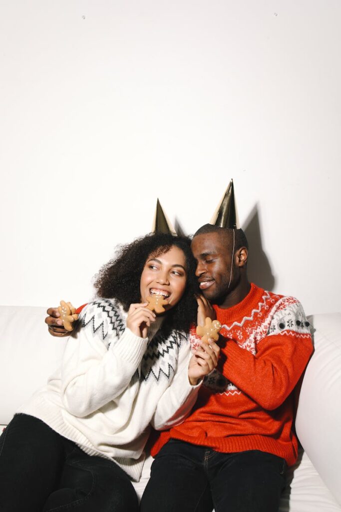 Happy couple in sweaters wearing party hats, enjoying gingerbread cookies indoors.