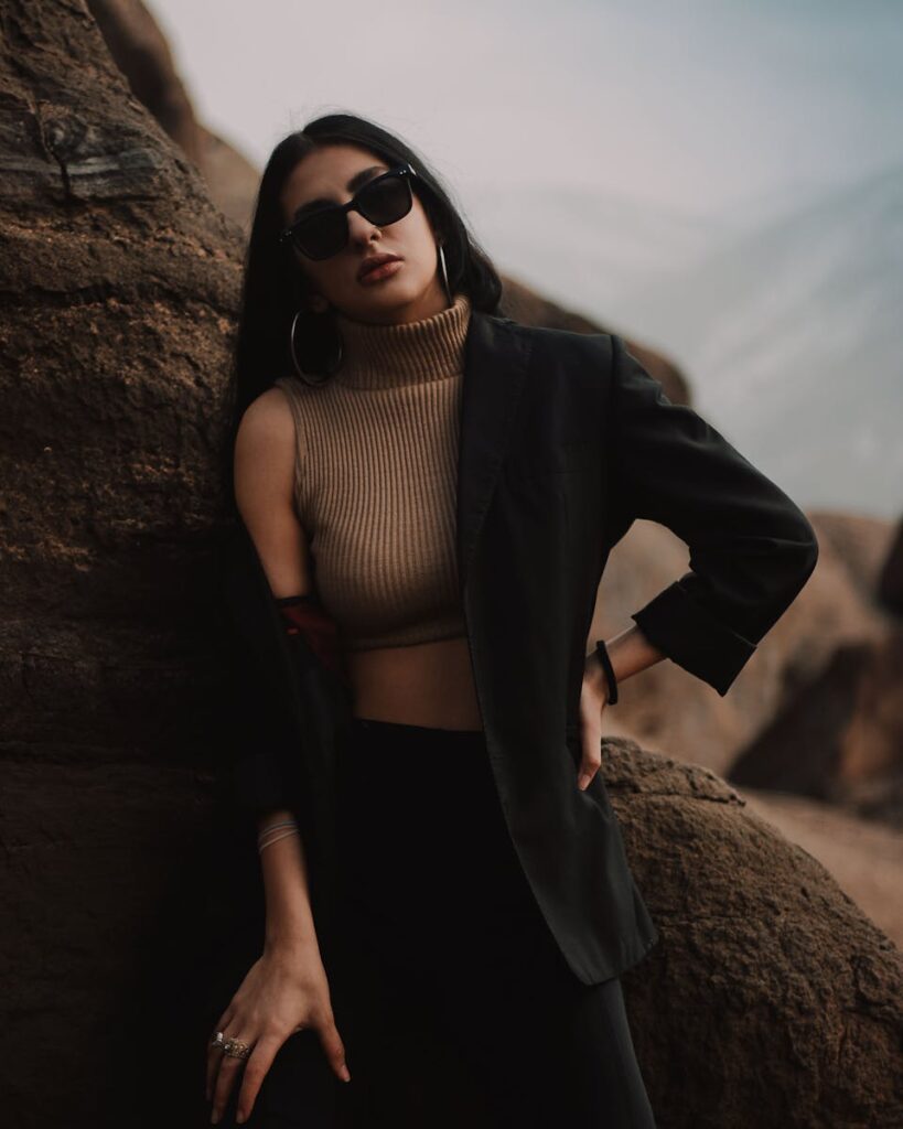 Stylish woman in sunglasses and blazer posing against rock formations in a moody setting.