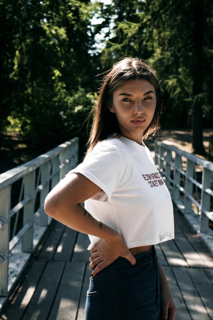 Portrait of a young woman posing confidently on a sunny wooden bridge surrounded by trees.