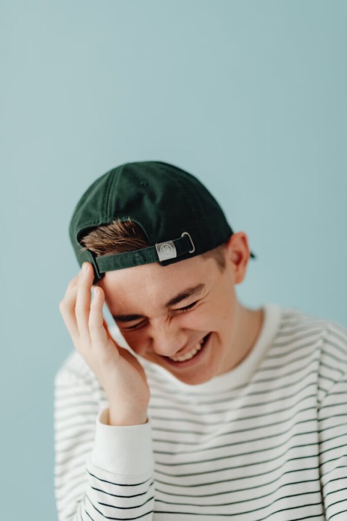 Playful portrait of a young man laughing while wearing a backward cap against a plain background.