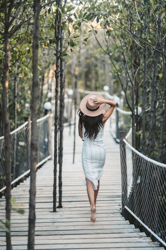 Elegant woman in a dress and sunhat walking barefoot on a scenic wooden bridge surrounded by nature.