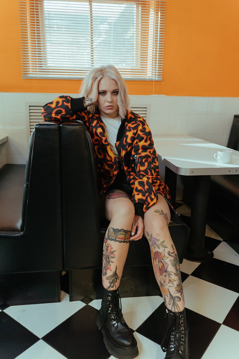 A tattooed woman with blonde hair in an orange jacket sits at a cafe booth looking serious.