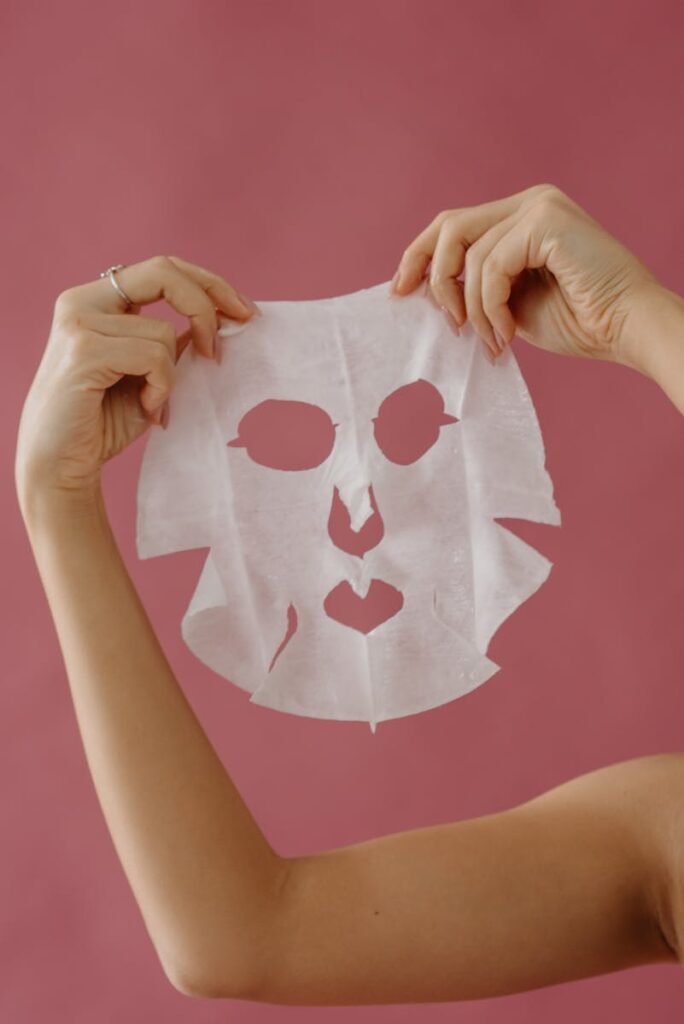 Close-up of hands displaying a sheet mask for skincare against a pink backdrop.