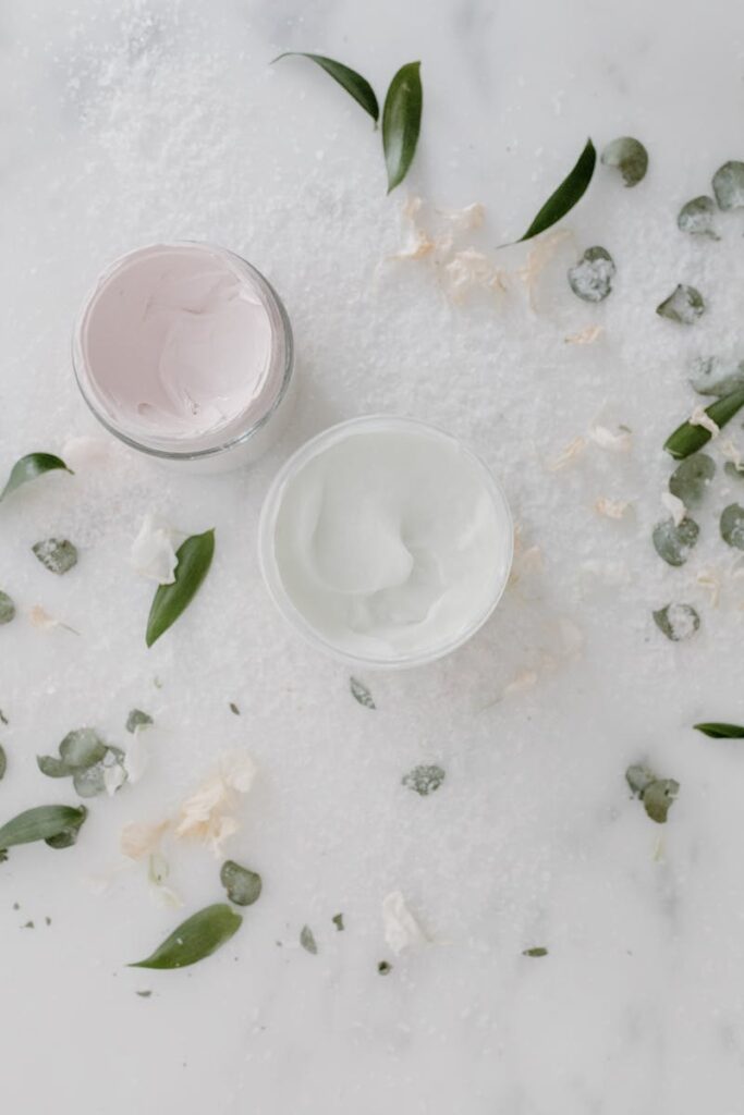 Overhead shot of cosmetic cream jars surrounded by green leaves on a marble surface.