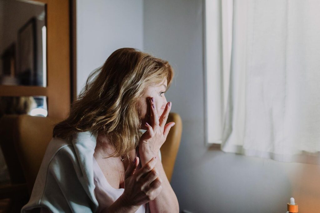 A woman applies skincare by a window, capturing a calm moment indoors.