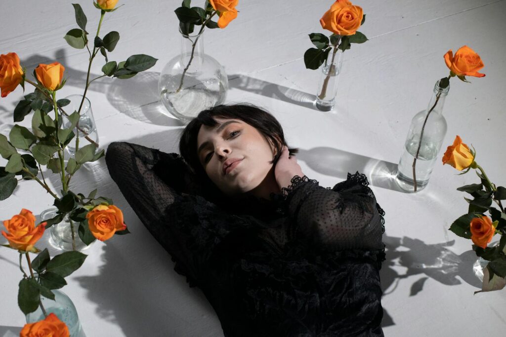 A woman in black lace lies among orange roses in delicate glass vases, creating a serene and stylish composition.