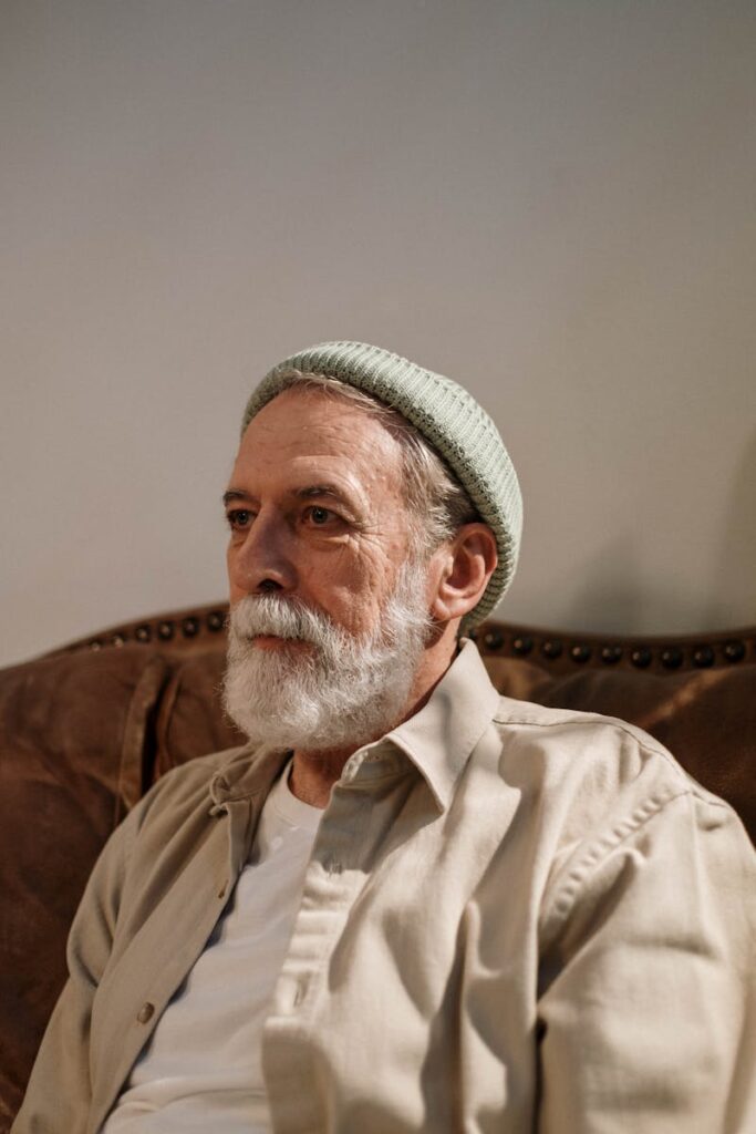 Elderly man with a serious expression wearing a beanie, sitting indoors on a couch.