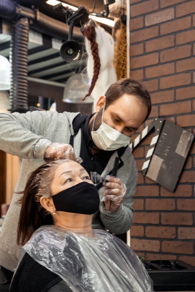 Bearded man in protective mask applying hair die for aged female in modern salon
