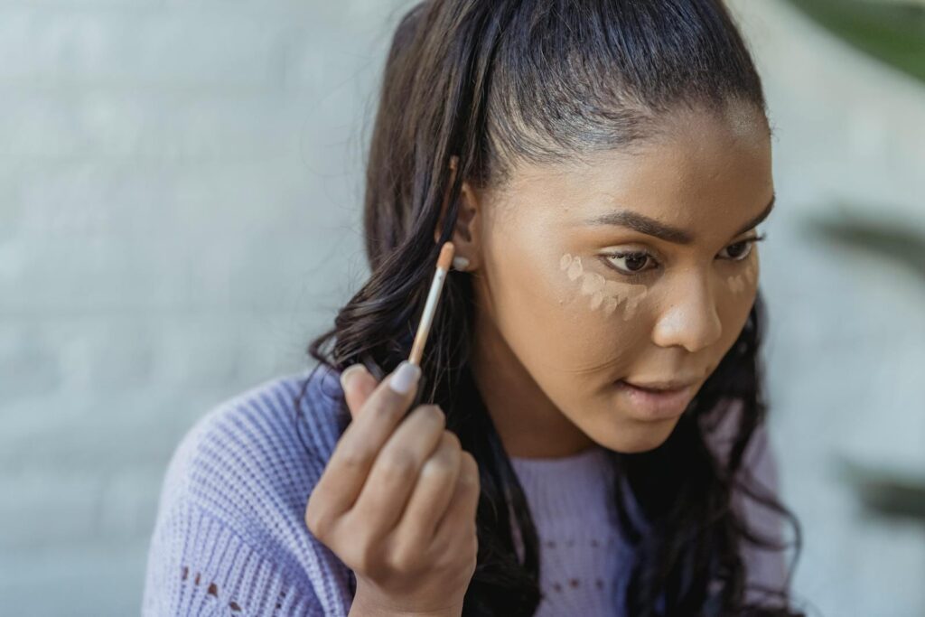Crop African American woman with long dark hair applying concealer under eyes and looking away in light room